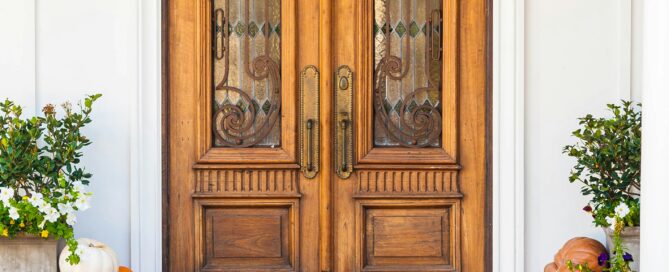 Arched front door with wood paneling and glass windows on a white house with orange and white pumpkins on the porch.